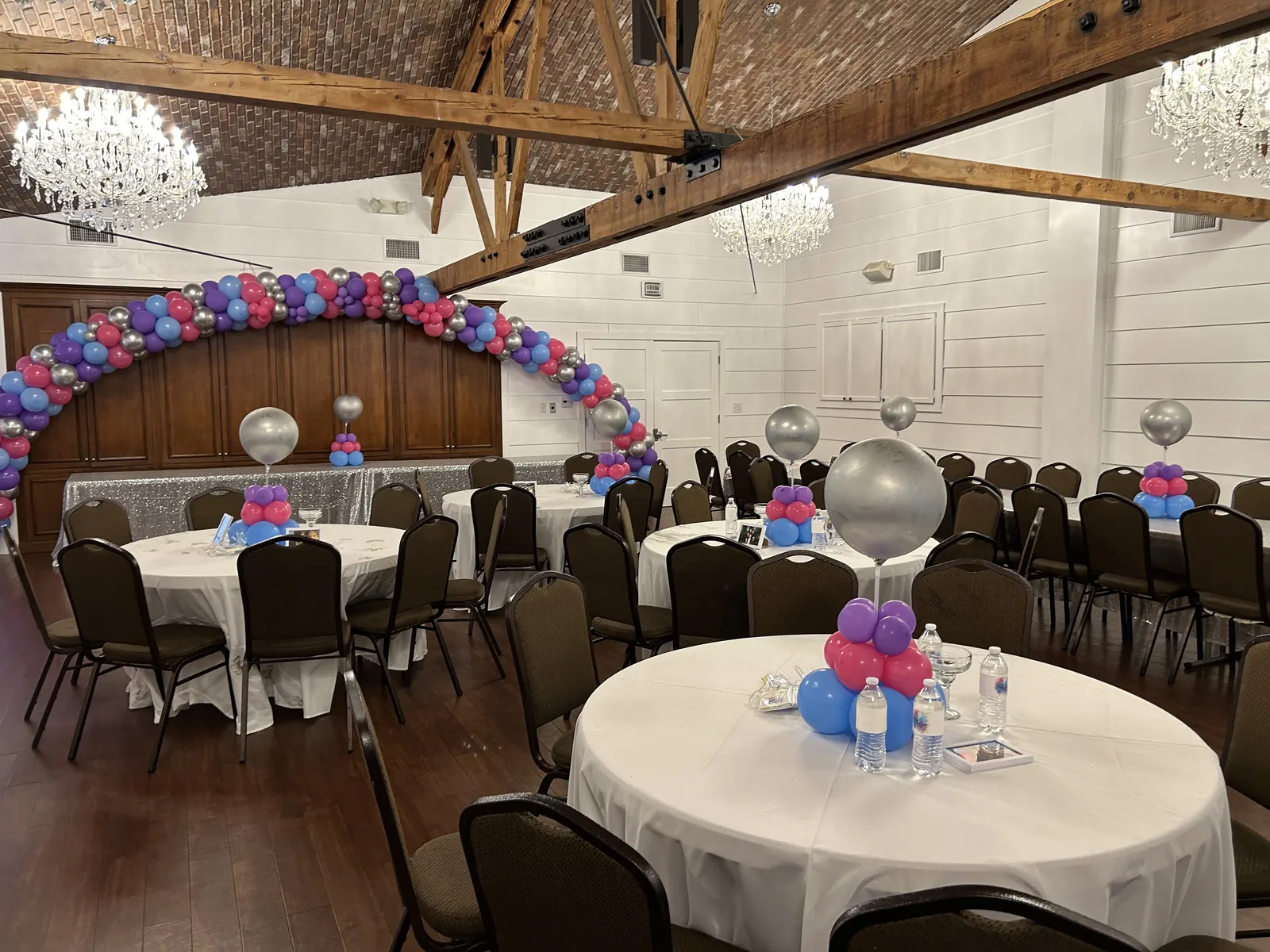 Extra large balloon arch in pink, purple and silver across an event hall with round tables and centerpieces