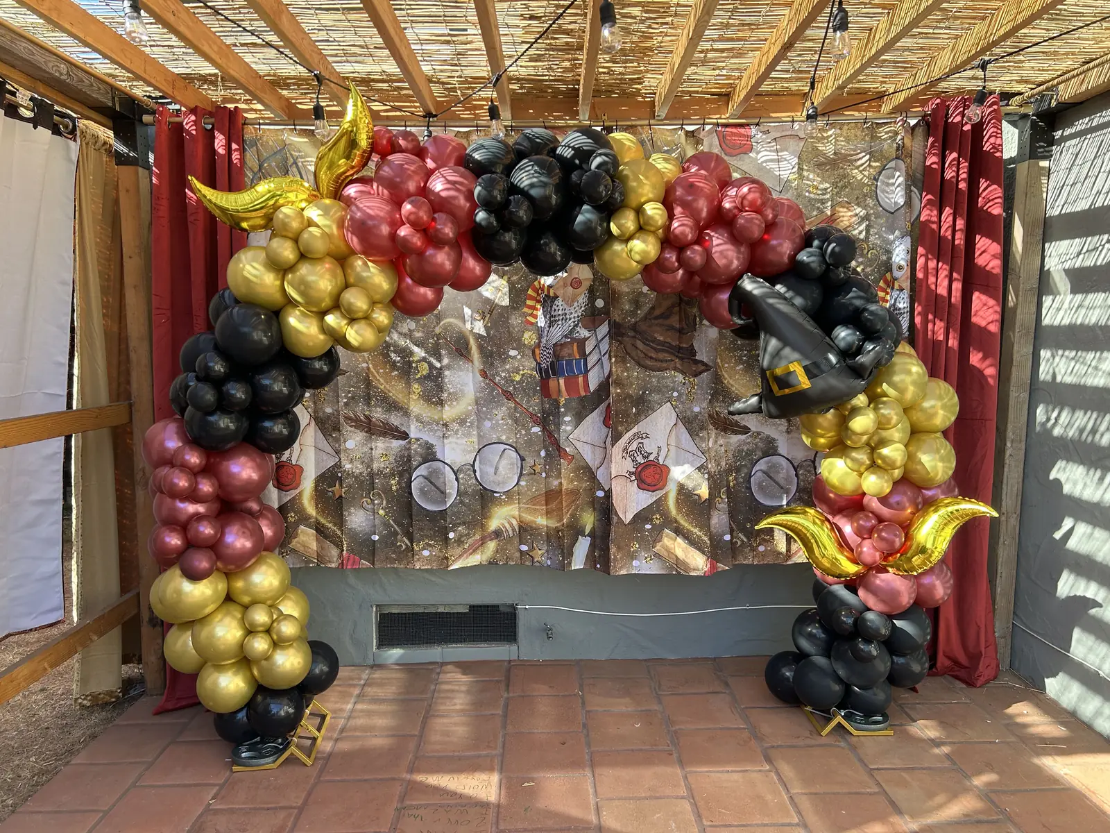 Burgundy, gold and black balloon arch with winged gold accents and a wizard hat