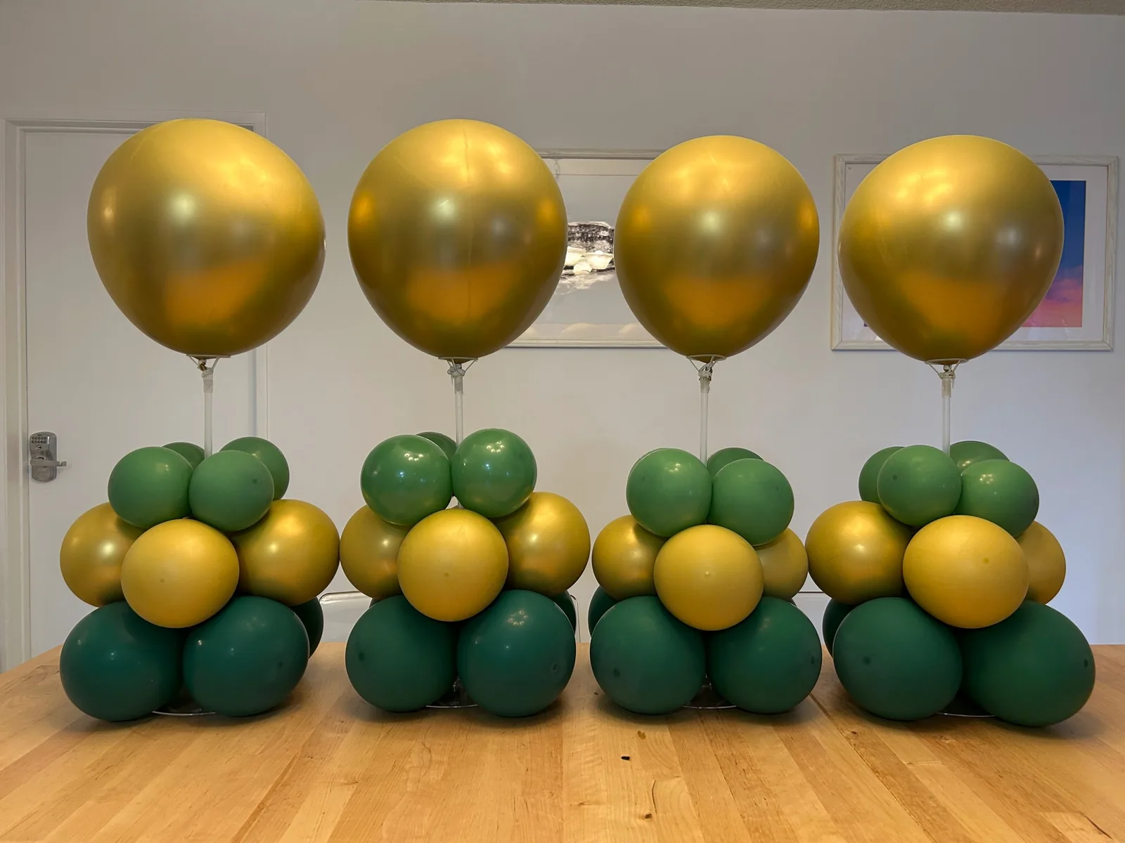 Four forest green, sage and chrome gold balloon centerpieces with gold topper balloons lined up on a table