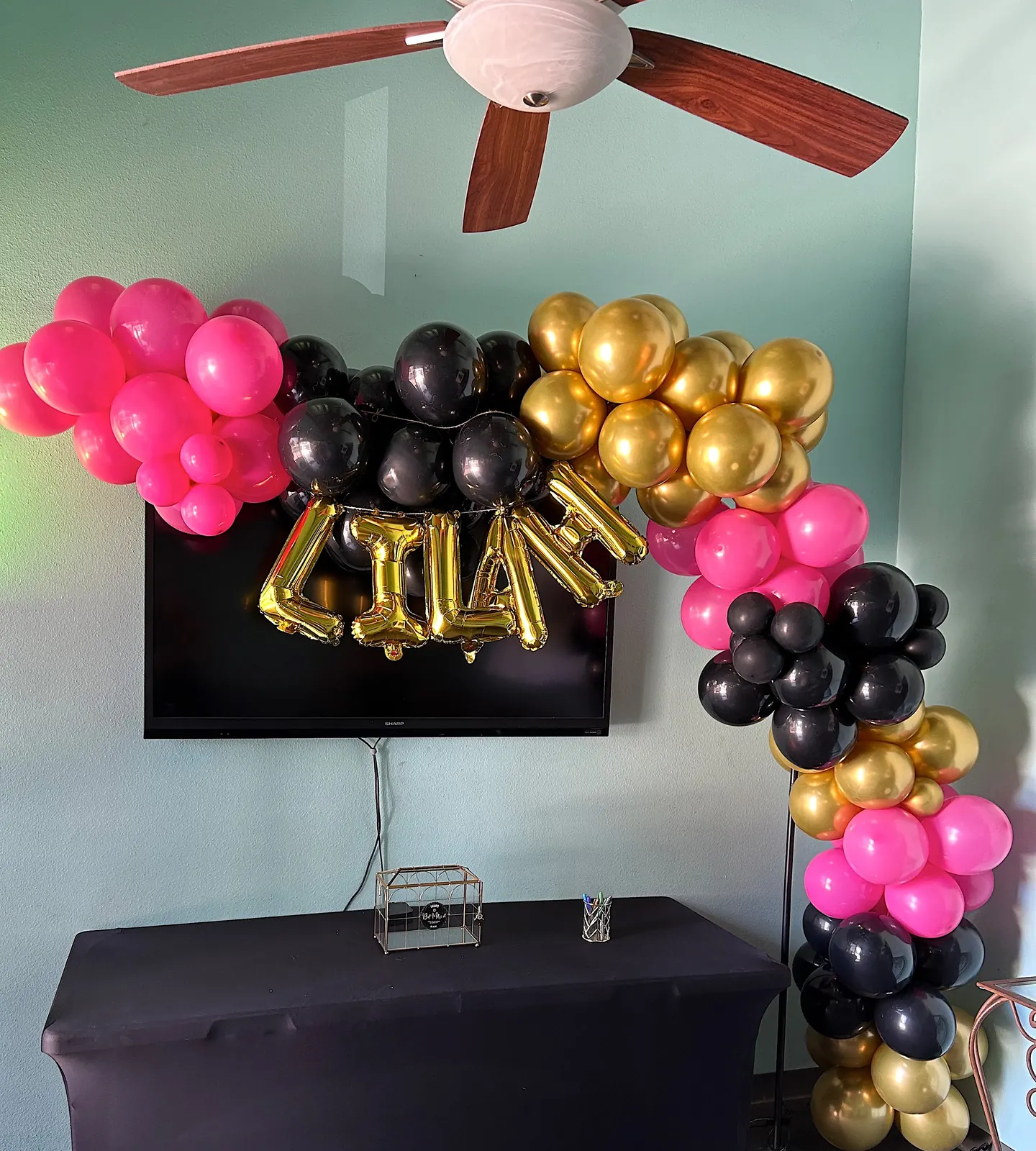 Hot pink, black and gold balloon garland around a TV wall with gold letter balloons