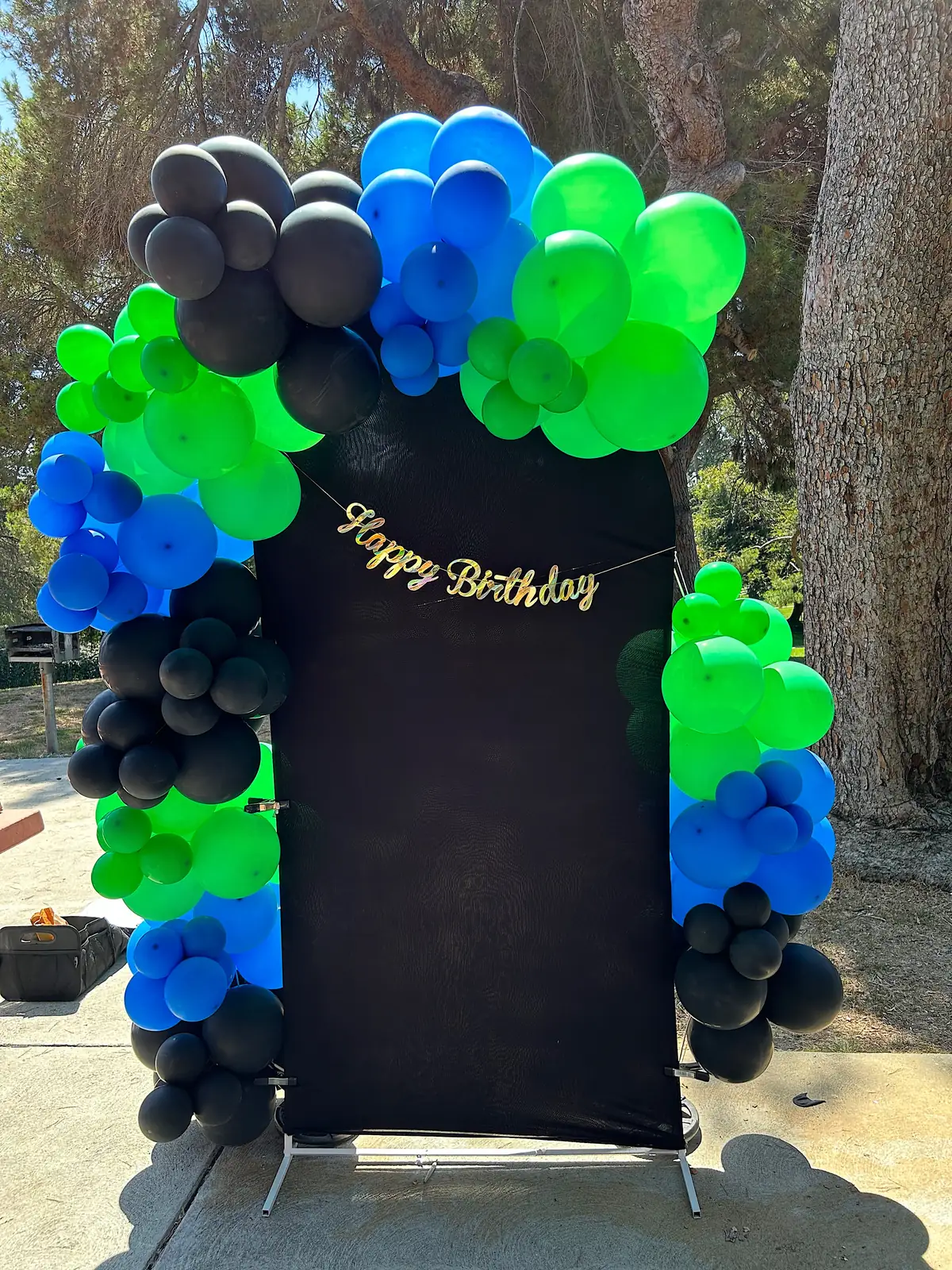Black Chiara backdrop with a Happy Birthday banner framed in green, blue and black balloons outdoors