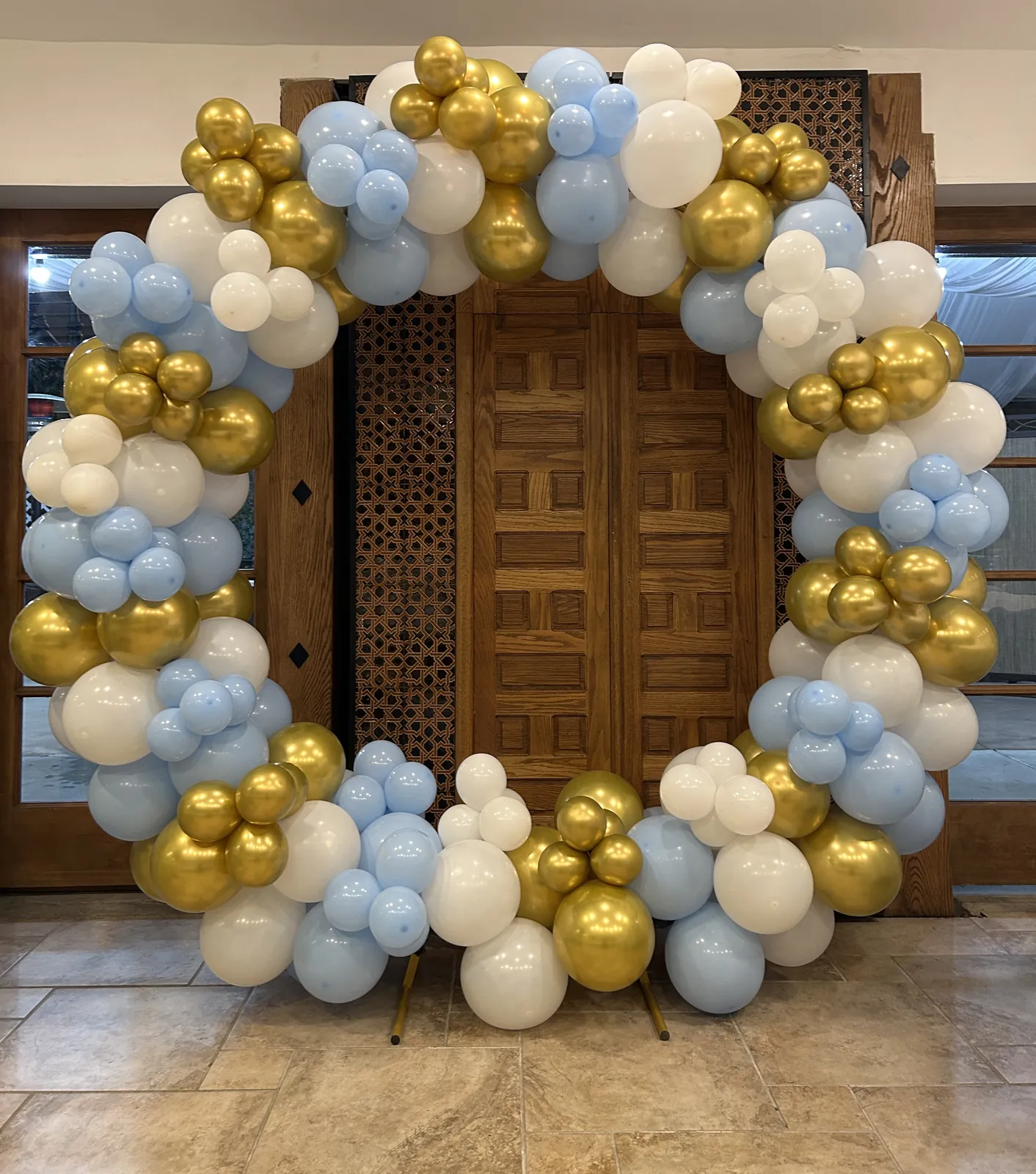 Circular balloon arch in baby blue, white and metallic gold in front of carved wooden doors for a bris in Beverly Grove, Los Angeles
