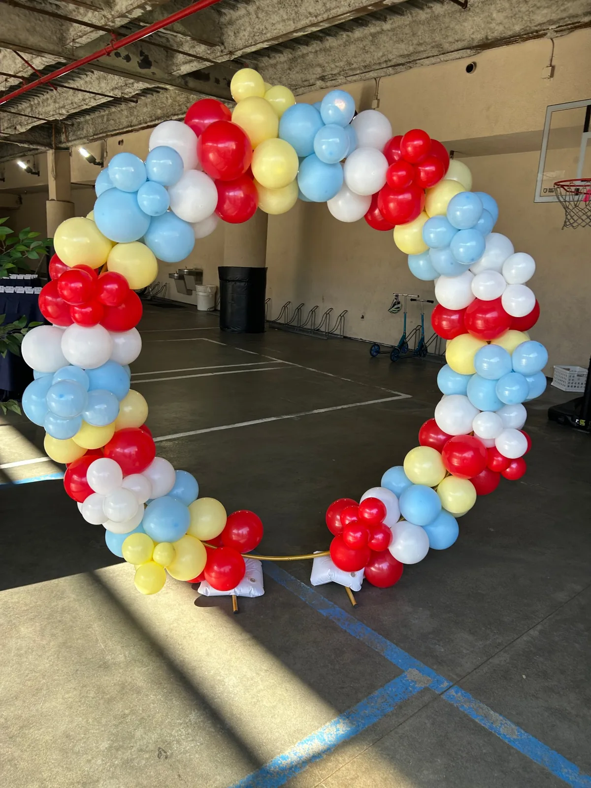 Red, light blue, yellow and white circular balloon arch on a gold ring stand in a covered court with a basketball hoop
