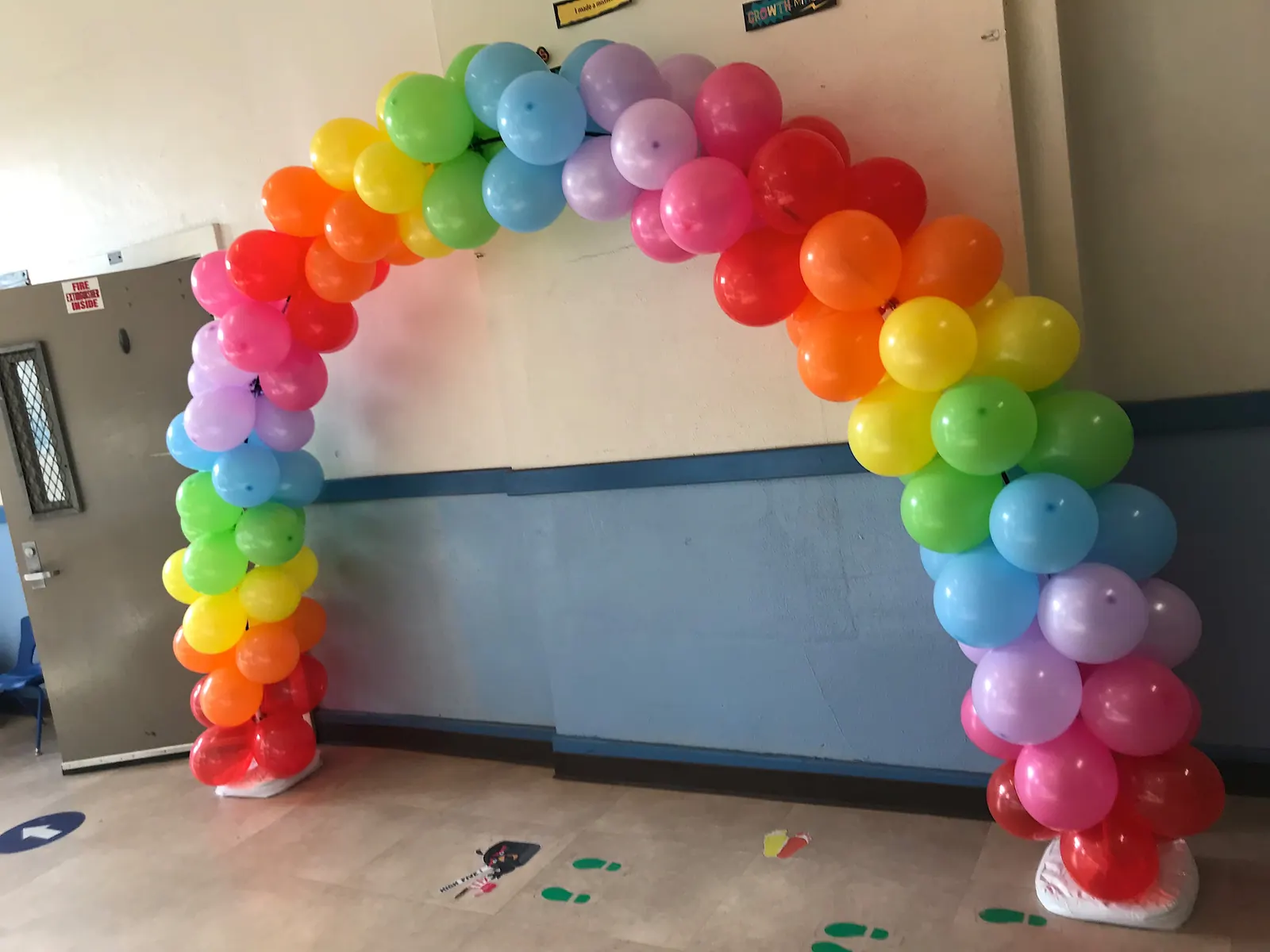 Rainbow table balloon arch on its own stands in a school hallway in Los Angeles